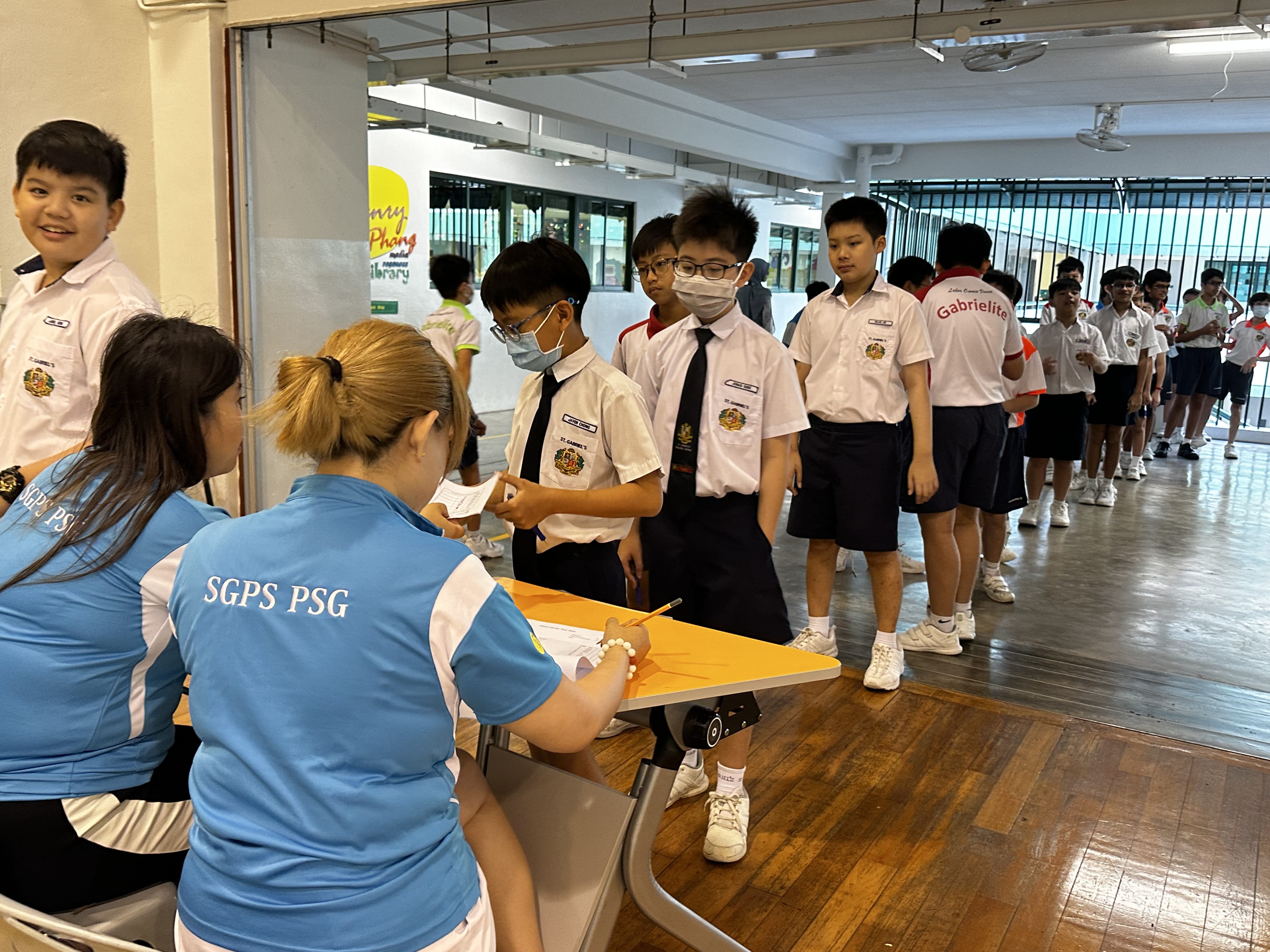 Students in school uniforms queue at a registration table staffed by two adults in blue SGPS PSG shirts.